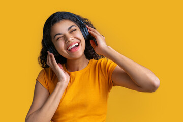 Wall Mural - Studio portrait of cute smiling casually dressed dark skinned girl in wireless headphones enjoying listening to music and singing isolated over bright colored yellow background