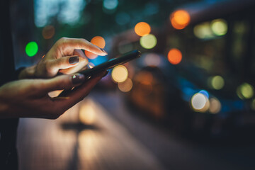 Wall Mural - Closeup female hands  with smartphone using application for calling taxi online walking on city street night, girl  with internet connection texting friend via mobile phone on background bokeh light