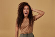 Stressed young brown haired curly dark skinned lady frowning her face while screaming and raising emotionally hand to her head while posing over beige background