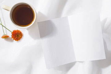 Top view of voile fabric background with a mug of tea, two orange flowers and a piece of white paper with space for text. Horizontal