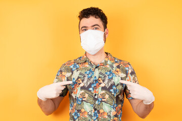 Wall Mural - Horizontal shot of cheerful stylish man points at his casual hawaiian shirt and medical mask and safety gloves, stands indoors against concrete wall, being in good mood after going shopping a