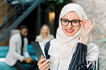 Wall Mural - Charming arabian Muslim business woman in white hijab holding folder of papers and posing on camera with happy smile, standing in modern loft office on the background of multiethnic colleagues