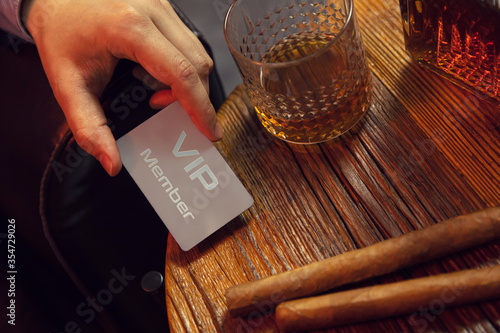 Man\'s hand puts VIP membership card on the table. Gentleman\'s hand puts exclusive VIP membership card on the wooden table with whisky in carafe and glass with cigars.