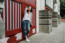 Full Length Of Young Asian Korean Woman Tourist With Bag Flicks Her Hair And Standing Against Red Door By Chinese Old Building. Happy Girl Traveler Relax Outdoor Waiting For Someone By Entrance.