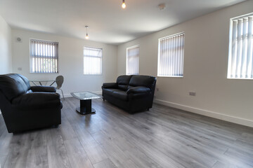 A modern British apartment living room and kitchen with black leather sofa and a coffee table on a grey wooden floor with white walls and modern fitted kitchen with a fridge