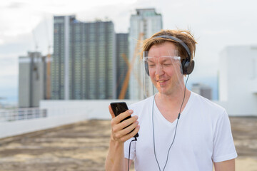 Poster - Happy young man with face shield using phone and listening to music at rooftop of the building