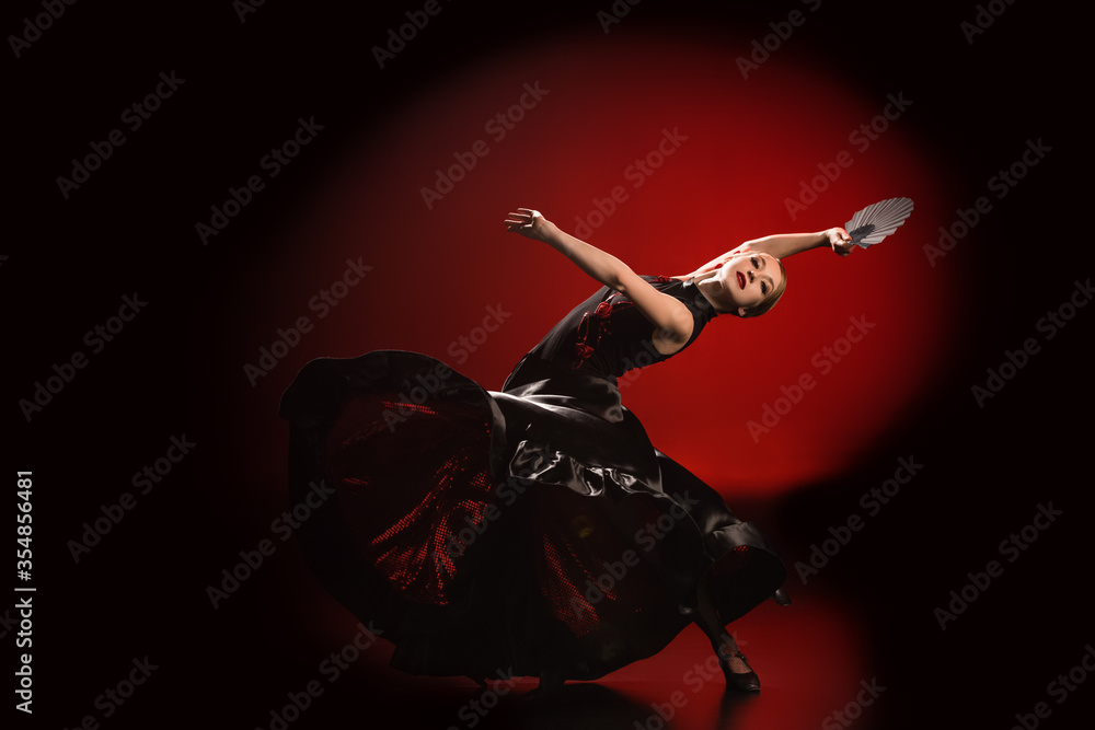 young flamenco dancer in dress holding fan while dancing on red and black - obrazy, fototapety, plakaty 