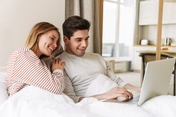 Poster - Portrait of joyful couple smiling and using laptop while lying in bed