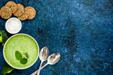 Wall Mural - Broccoli cream soup, bread, spoon on blue kitchen desk top-down space for text