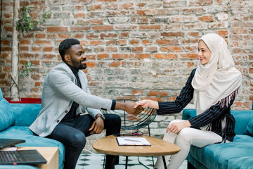 Wall Mural - Two confident business people, African man and Muslim woman in white hijab, shaking hands during a meeting in the office, after successful dealing, signing the contract, greeting and partner concept.