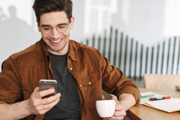 Image of joyful man smiling and using cellphone while drinking coffee