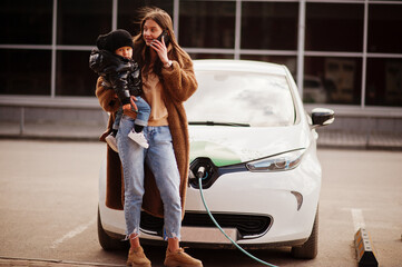 Wall Mural - Young mother with child charging electro car at the electric gas station and speak on mobile phone.