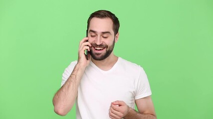 Wall Mural - Bearded happy smiling young guy in white t-shirt using speaking talking on mobile cell phone conducting pleasant conversation isolated on green chroma key background studio. People lifestyle concept