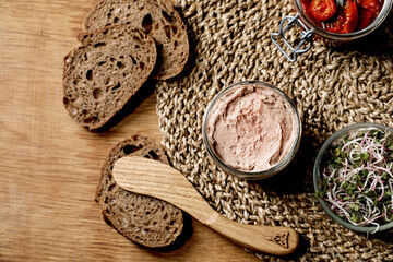 Wall Mural - Glass jar of homemade chicken liver pate with wooden knife, sliced rye bread, sun-dried tomatoes and green sprout salad on wooden background. Home breakfast or appetizer. Flat lay