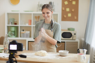 Smiling young woman making dough while standing in domestic kitchen and shooting the video for her blog