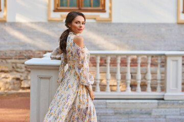 Wall Mural - Pretty smiling young woman with long brunette hair wearing fashionable floral dress standing on street against balustrade on background. Happy female model in elegant stylish outfit posing outdoors.