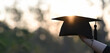 © Prathankarnpap - Cropped image of university student hand holding a graduation hat in hand over outdoors with sunset as background.
