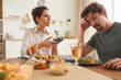 © AnnaStills - Young woman pointing at mobile phone and talking to man during dinner in the kitchen at home
