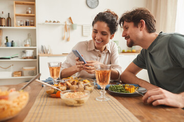 Young smiling woman showing some pictures on her mobile phone to her boyfriend during dinner in the kitchen at home