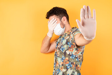 Wall Mural - People, body language. Young man wearing hawaiian shirt and medical mask covers eyes with palm and doing stop gesture, tries to hide from everybody. Don't look at me, I don't want to see, 