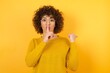© Roquillo - Young beautiful African American woman with curly hair wearing yellow sweater asking to be quiet with finger on lips pointing with hand to the side. Silence and secret concept.