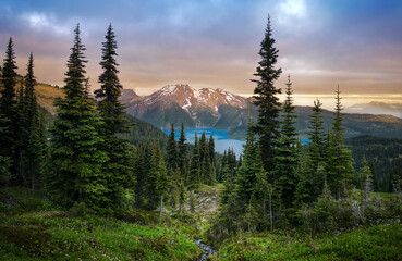 Wall Mural - Glacial mountain Garibaldi lake with turquoise water in the middle of coniferous forest at sunset. View of a mountain lake between fir trees. Mountain peaks above the lake lit by sunset rays. Canada