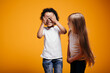 Long-haired girl child checks whether a dark-skinned boy peeps when playing hide and seek on an orange background in the studio.
