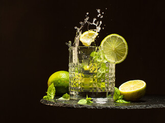 Wall Mural - Glass of Mojito cocktail with fresh limes,mint leaves,ice cubes and refreshing water.Splash of cocktail Mojito on black stone plate.