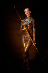 Valkyrie girl in shiny military armor and with a spear in a dark room with plants and vines. Model during a photo shoot, the actress during the shooting of the film.