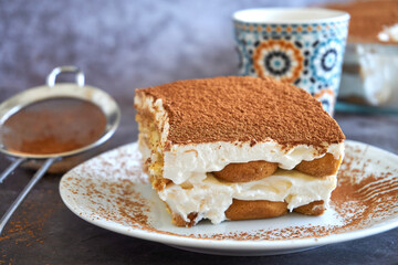 Square slice of tiramisu cake and a cup of coffee in the background, homemade gluten-free Italian layered dessert with ladyfinger biscuit and mascarpone cream decorated with cocoa powder