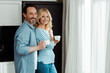© LIGHTFIELD STUDIOS - Side view of mature couple smiling at camera while holding coffee cups in kitchen