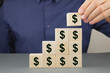 © VITAL - Closeup of a businessman making a pyramid of wooden cubes with a dollar sign on them. Financial planning concept.