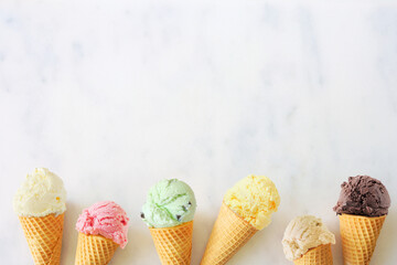Wall Mural - Ice cream cone bottom border with a variety of flavors. Overhead view on a white marble background with copy space.