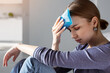 Woman applying ice pack as cold compress on forehead due to headache, migraine, tired after work, sitting indoors in apartment. Put cold on head for relief hurt and fever