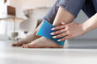 Close up woman feet and legs, and hands holding ice gel pack as cold compress on ankle due to stretching or injury, sitting on floor in apartment indoors
