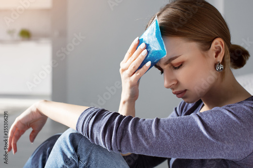 Woman applying ice pack as cold compress on forehead due to headache, migraine, tired after work, sitting indoors in apartment. Put cold on head for relief hurt and fever
