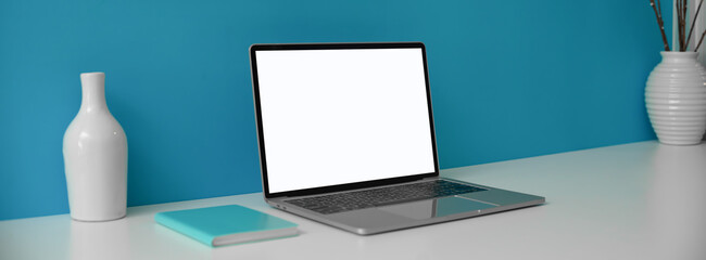 Creative workspace with mock-up laptop, notebook and ceramic vases on white desk with blue wall