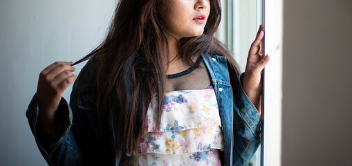 Wall Mural - Half face portrait of an Indian Bengali beautiful and cute brunette girl in a casual blue jeans shirt and white top standing near a window. Indian lifestyle and fashion portrait