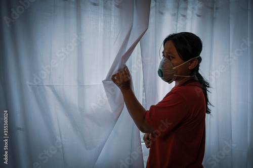 Cuadros en Lienzo A woman with face mask on anxiously peeks through her window curtains anticipating dangers coming her way