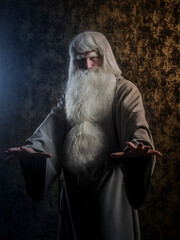 Wall Mural - A grey-haired bearded wizard in a gray cloak is practicing sorcery against a dark textured golden background.