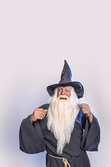 Wall Mural - A severe grey-haired bearded sorcerer in a gray cassock and a cap is practicing sorcery and doing magic against a light background.