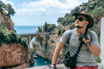 Sticker - The handsome man traveler enjoys a wonderful view of the beach and the stone bridge. Sunglasses and a kangaroo leather cowboy hat. Travel and vacation. Fiordo di Furore, Positano