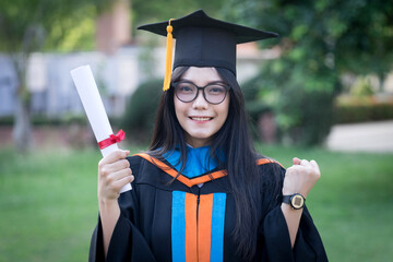 Wall Mural - Portrait of happy and excited of young Asian female university graduate wears graduation gown and hat celebrates with degree in university campus in the commencement day. Education concept.
