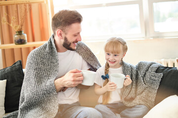 Father and daughter. Handsome young man and little cute girl sitting on the sofa at home, covered themselves with a warm blanket, have fun, holding white cups. Dad and child laugh. Father's day.