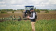 © DiedovStock - Bearded agronomist wearing a cap uses a specialized app on a digital tablet PC on the background of working tractor with a cultivator in the field