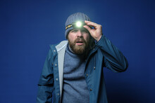 Bearded Man With A Headlamp In A Knitted Hat And A Waterproof Jacket Is Trying To See Something In The Light Of A Flashlight.