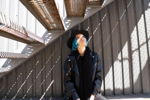 A Girl With Blue Hair Sits Under A Metal Staircase Near A Gray Wall Of Metal Siding