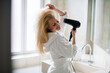 © JustLife - Young happy smiling woman wearing bathrobe and  using hair dryer in bathroom. Beautiful woman enjoying in hotel room.