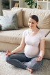© pressmaster - Young positive pregnant woman in activewear sitting in pose of lotus by couch