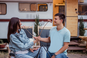 Happy young family at a campsite fries sausages on a bonfire. Picnic of a beautiful couple in love at the trailer, sitting comfortably, hugging, battle on sausages, kissing, laughing. Rest in nature